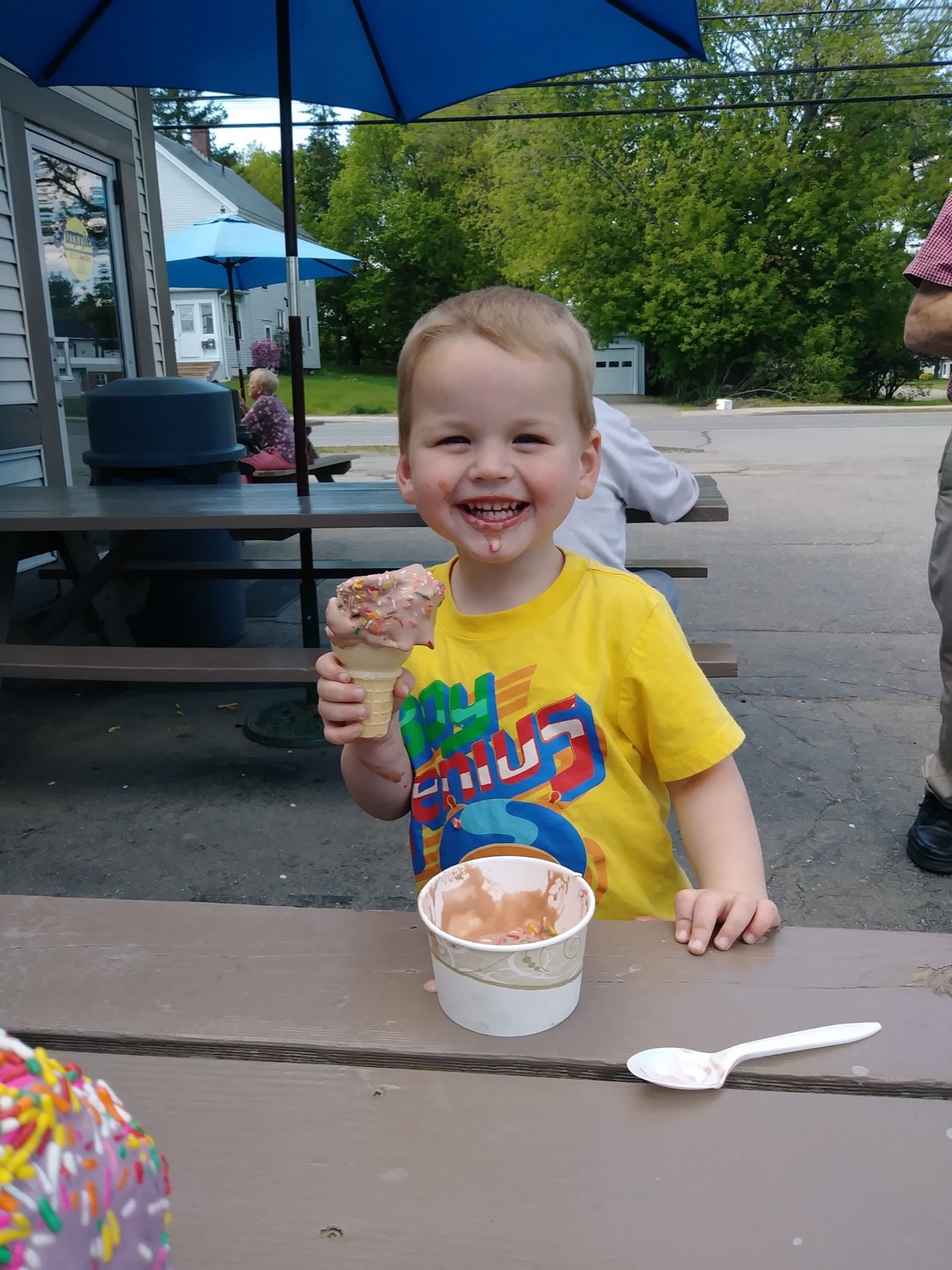 A smiling young boy at a picnic table holding a soft ice cream cone.