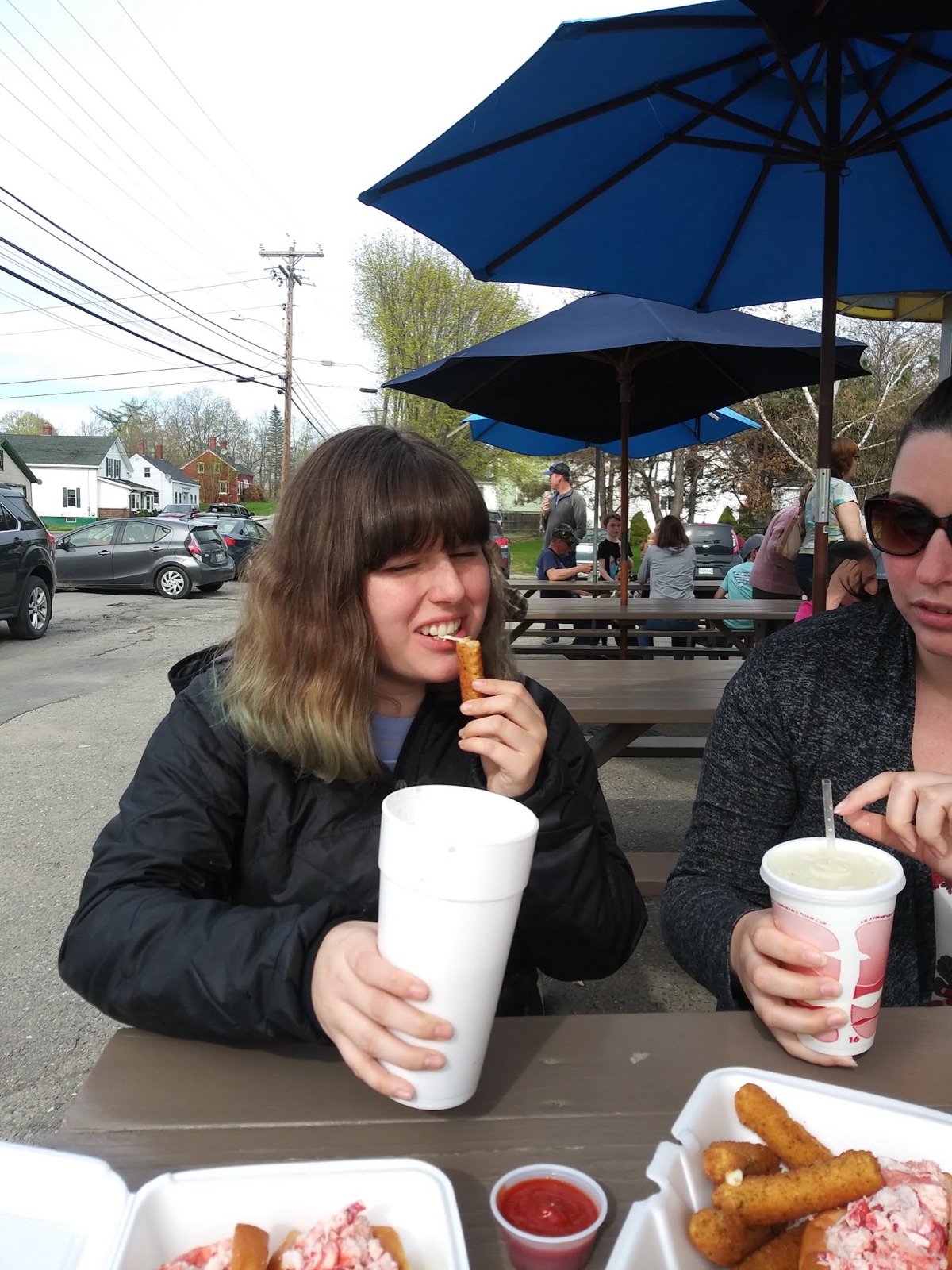 Two people eating at a picnic table under blue umbrellas outside the shop on a spring day.